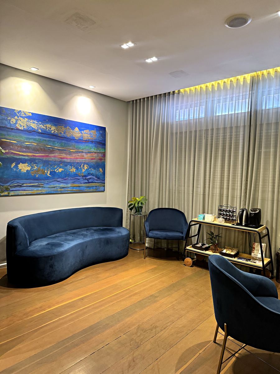 Recepção do primeiro piso da Santé Consultórios: Sala ampla com piso de madeira, cortina de parede inteira e luz indireta — hoje montada como espera, com sofá, poltronas e apoio de café.